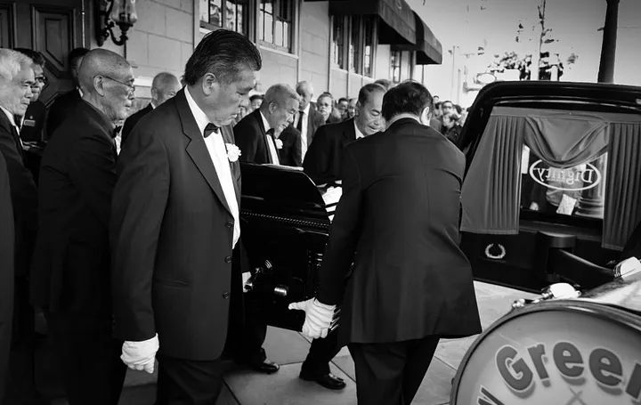 Pallbearers in formal attire with white gloves carrying a casket during a traditional Asian funeral procession with Vietnamese and Chinese ceremonial elements led by Green Street Memorial Band (formerly Green Street Mortuary Band)